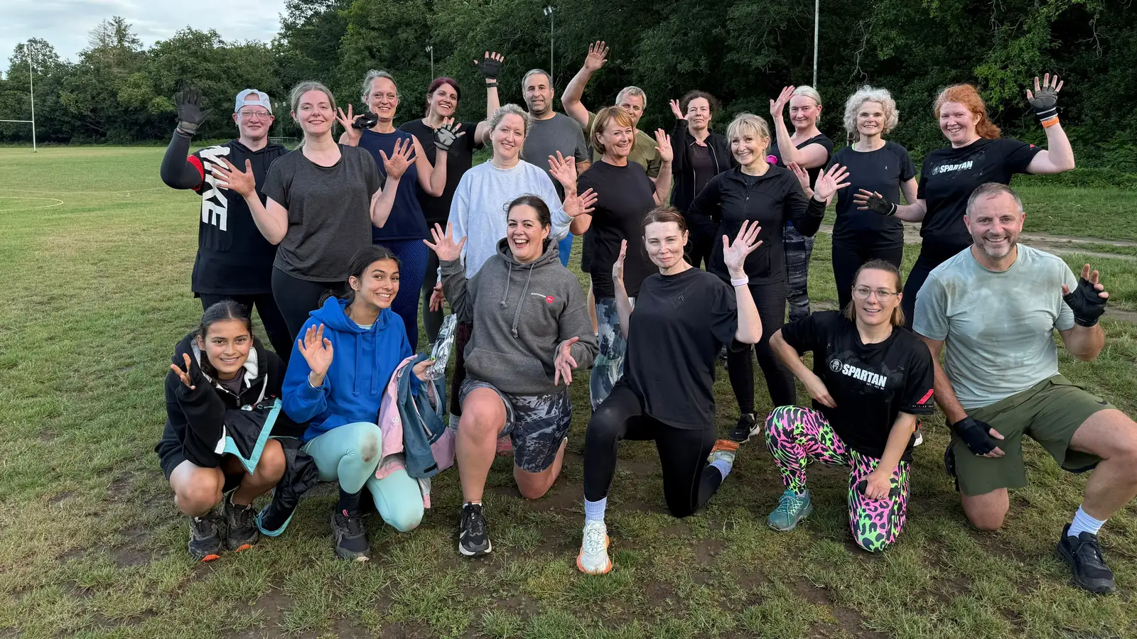 A mixed Bootcamp BR2 group waving on the rugby pitch in Bromley, with the posts and floodlights behind them
