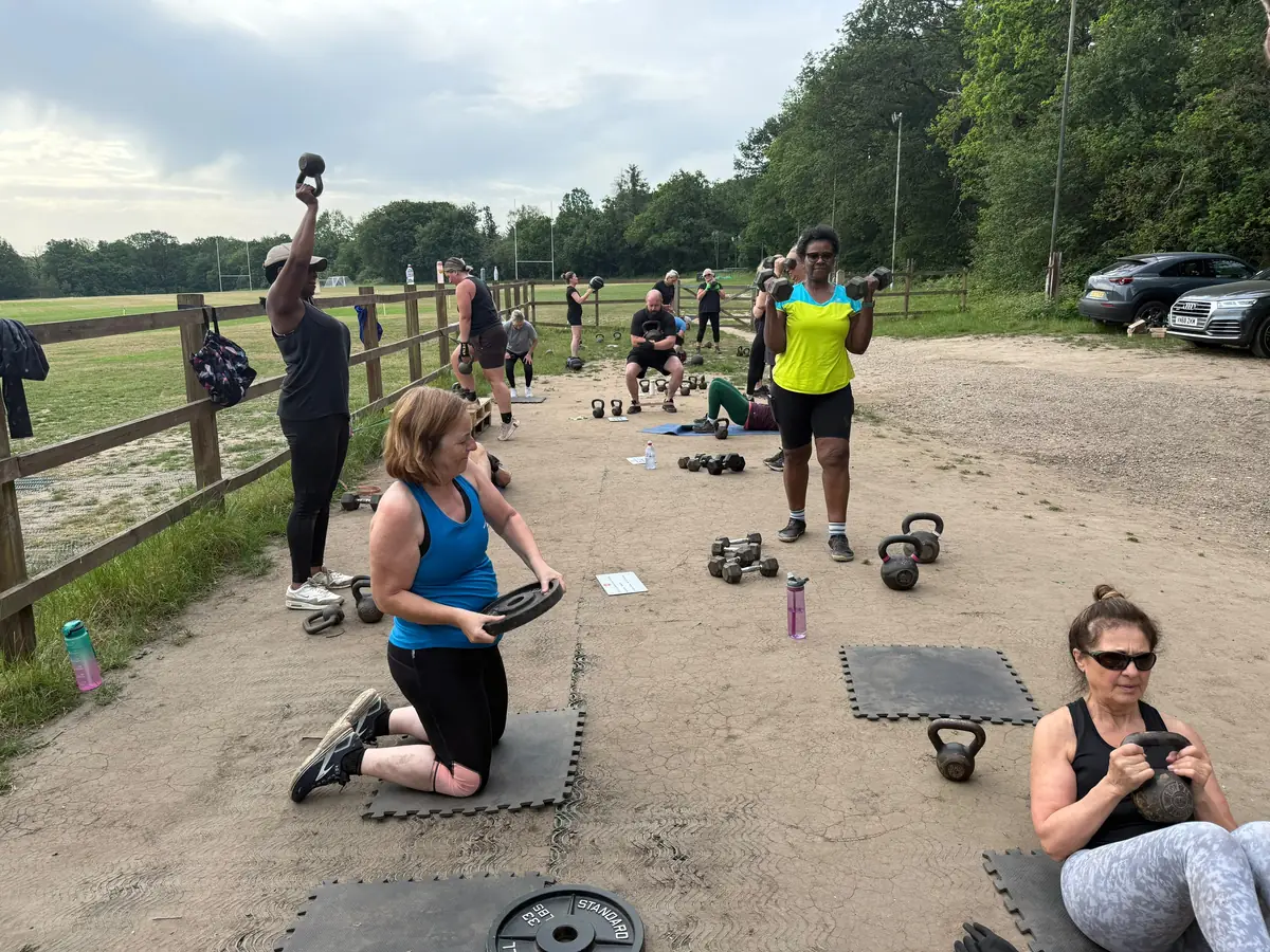 A Bootcamp BR2 session in progress by the fence at the rugby club, members working with kettlebells, dumbbells and weight plates