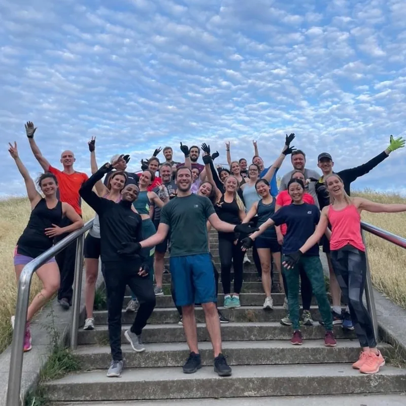 A Bootcamp SE16 group with arms raised on the steps at Stave Hill under a bright blue sky