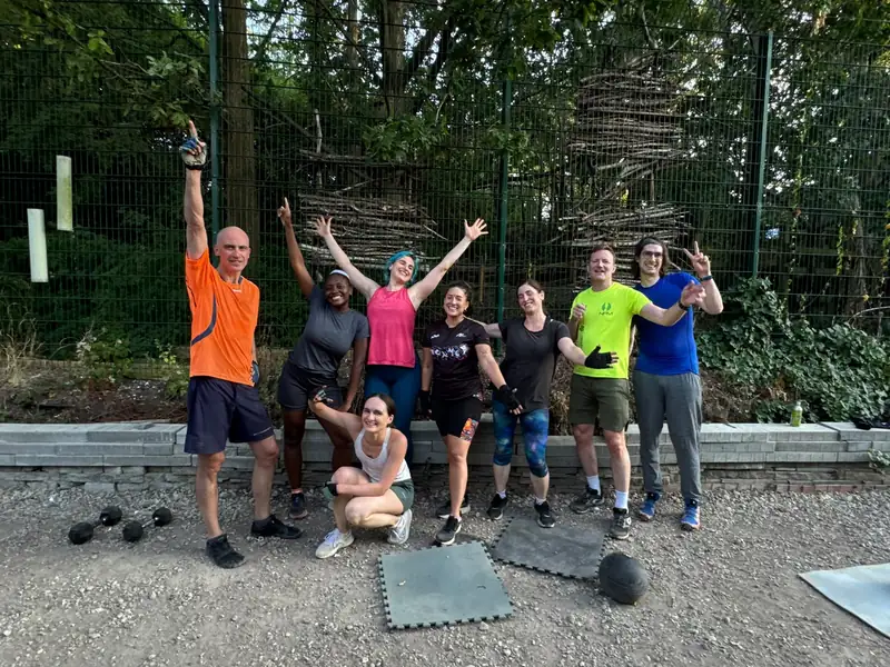A mixed group of Bootcamp SE16 members cheering by the shed after a session, kettlebells and mats around them