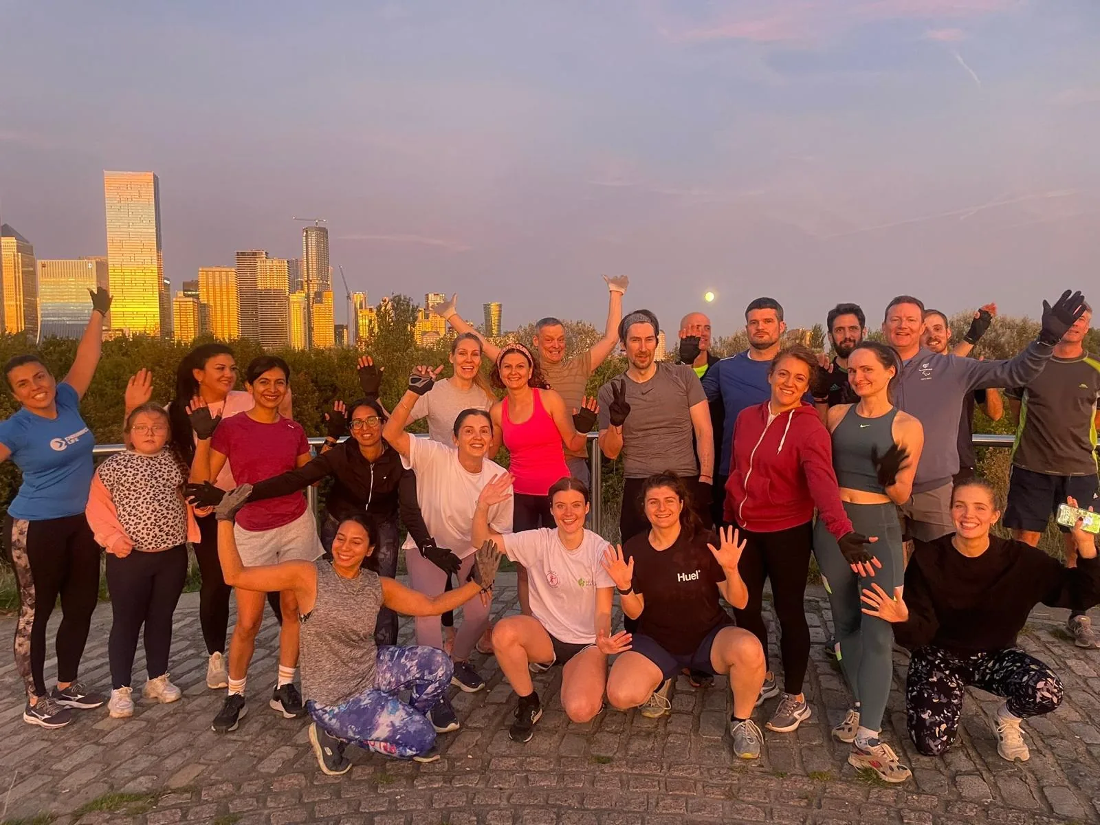 A large Bootcamp SE16 group smiling with their arms raised on top of Stave Hill, the London skyline lit up behind them