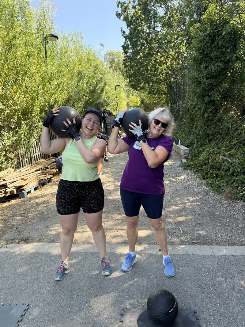 Two members smiling with medicine balls held up on their shoulders