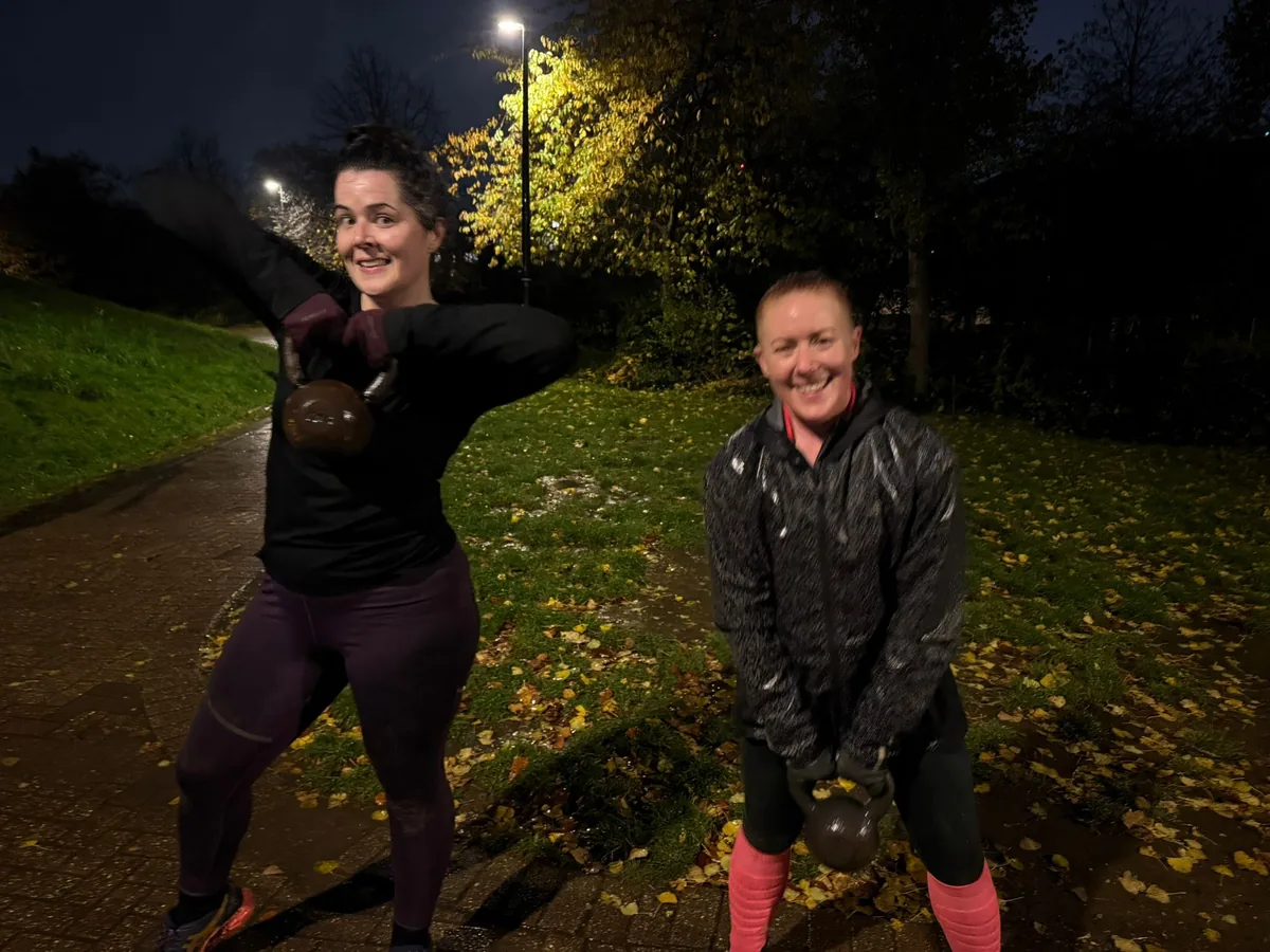 Two Bootcamp SE16 members smiling with kettlebells at an evening session in the dark, autumn leaves on the ground