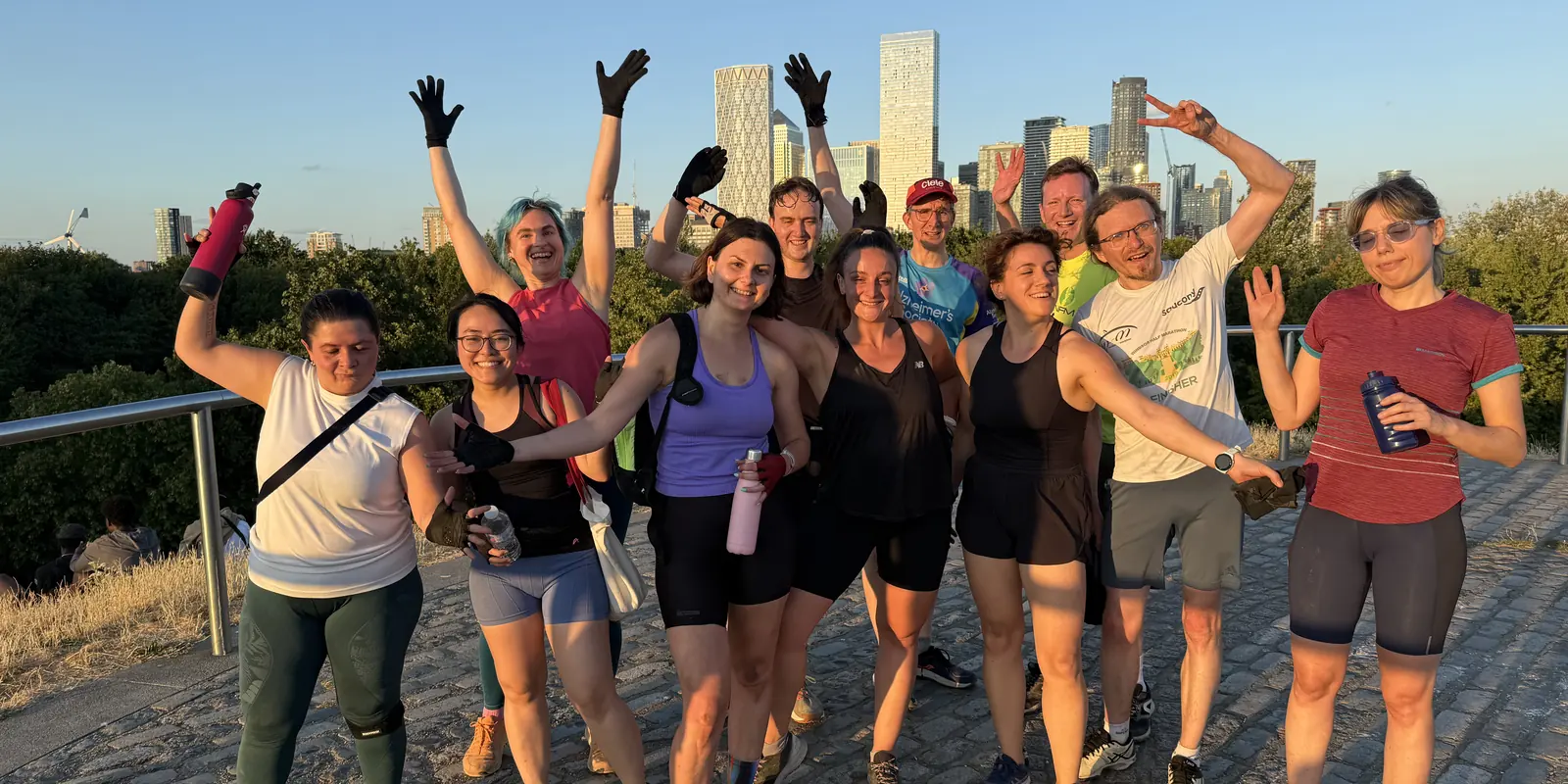 A large Bootcamp SE16 group with their arms in the air at Stave Hill, the Canary Wharf skyline behind them in the evening sun